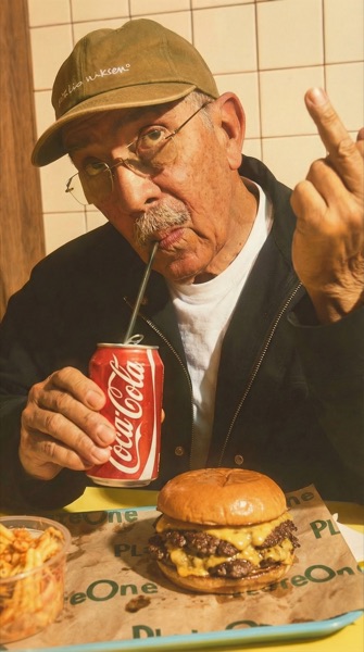 Un homme attablé devant un burger et des frites, une casquette sur la tête.